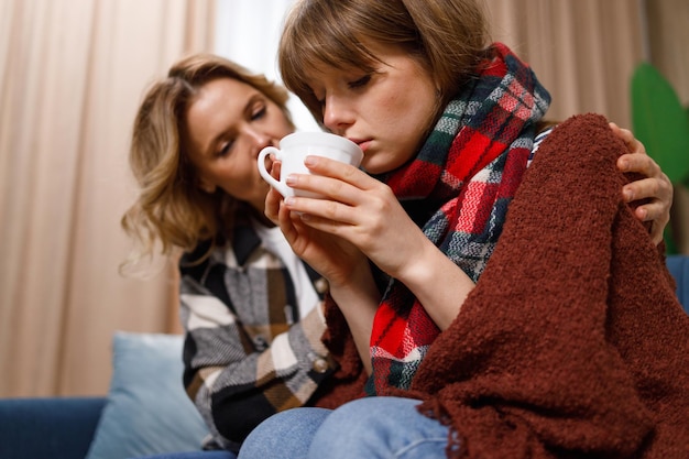 Caring woman hugging her daughter who is sick with the flu Ill girl in a blanket drinks hot tea