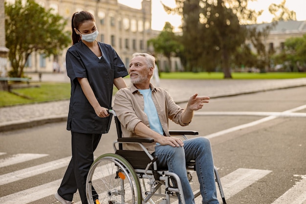 Caring young nurse in protective face mask having a conversation with mature disabled man in