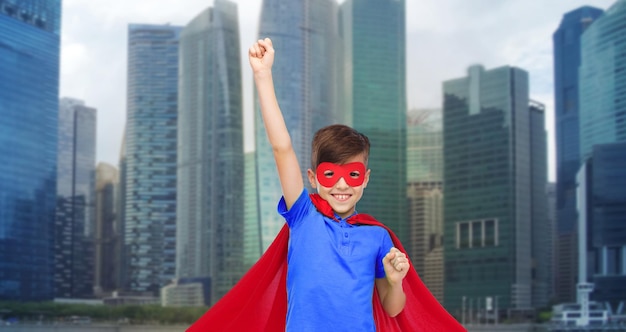 carnival, childhood, power, gesture and people concept - happy boy in red super hero cape and mask showing fists over city background