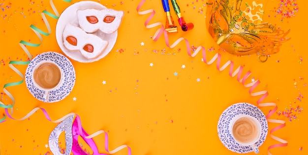 Carnival mask, two cups of aromatic coffee, sweets and confetti on a yellow background. Decor for a traditional Italian holiday. Copy space. Flat lay