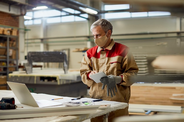 Carpenter with face mask using laptop and working on touchpad in a workshop