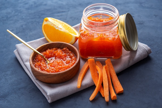 Carrot jam in a jar and a wooden bowl against a dark surface