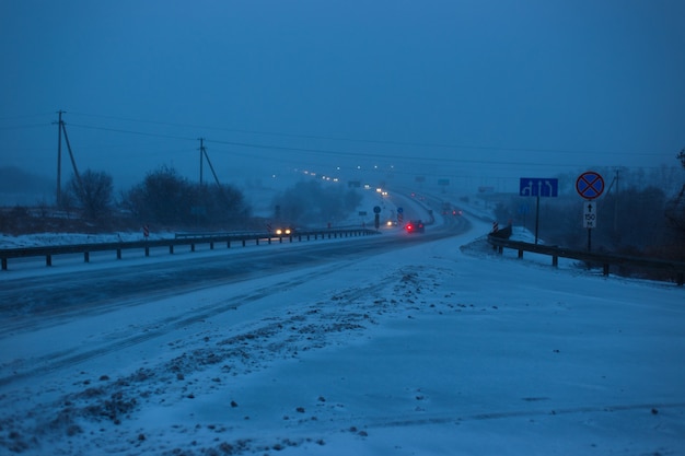 Cars drive on the highway during a snowstorm in the evening.