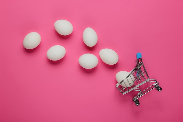 Cart for shopping and chicken eggs on pink