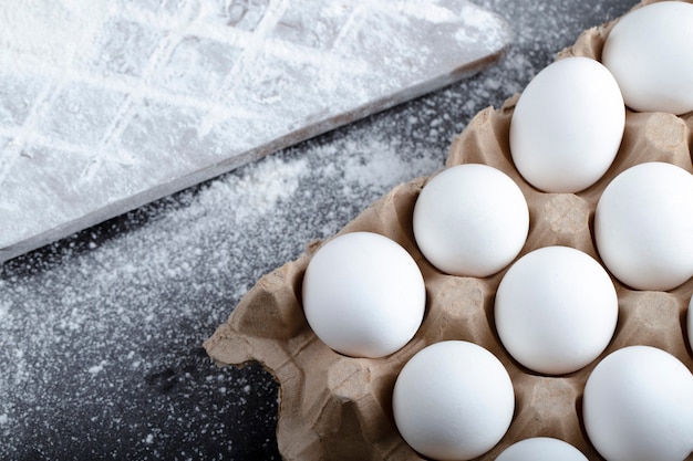 Carton container of raw eggs and spilled flour on black surface.