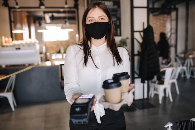 Cashier in face mask holding credit card reader machine and two cups of coffee contactless payments