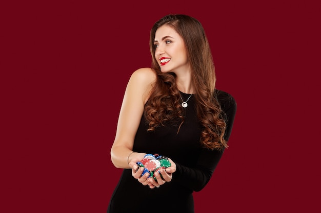 Photo casino, gambling, poker, people and entertainment concept - woman poker player in black dress with chips in hands on red background.