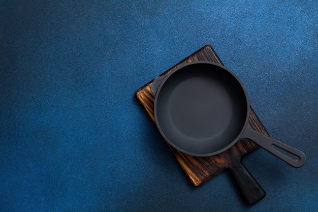 Cast iron skillet on a wooden board. Frying pan and cutting board on a dark blue background. Kitchen utensils. Empty place. Top view.