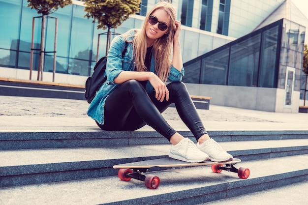 Casual blond female in denim shirt posing on stairs with longboard over glass building.