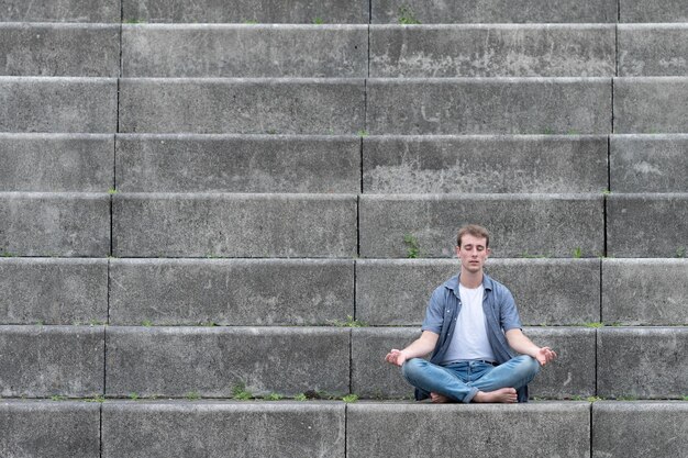 Casual dressed young man sitting on stairs and meditating. Mental health, mindfulness and yoga concept with copy space.