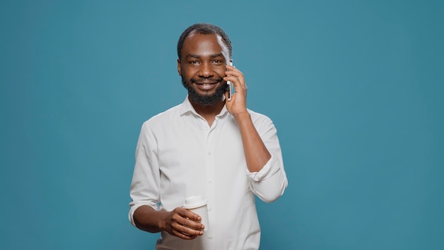 Casual entrepreneur with coffee cup talking on phone call in front of camera, enjoying lunch break. Man using smartphone to have conversation online, chatting about freelance work.