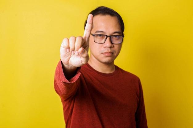 Casual man in glasses raises index finger warning gesture ban isolated on yellow background