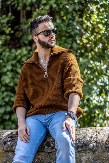 A casual man wearing a brown sweater with sunglasses looking away sitting on a stone