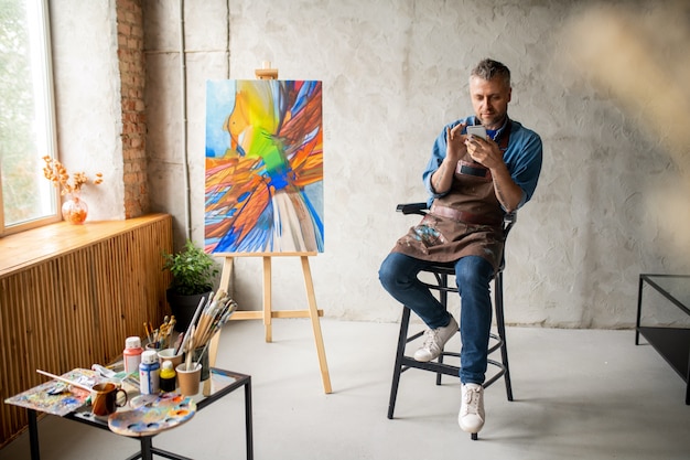 Casual painter with smartphone sitting in studio and scrolling in mobile phone