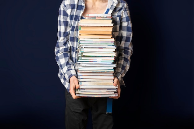 Casual student carry huge stack of books on dark background f