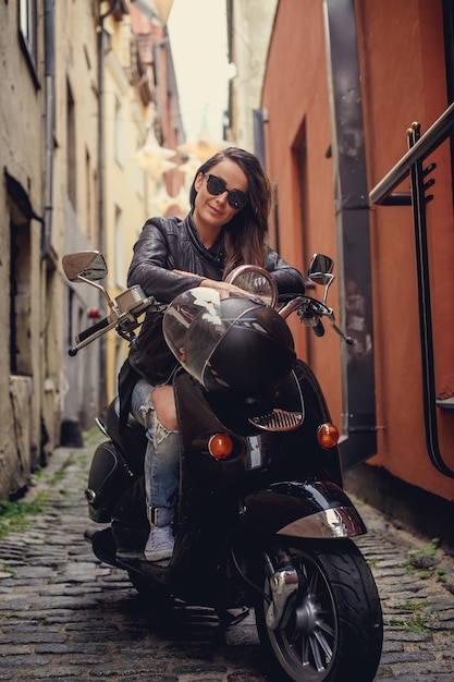 Casual woman with long brown hair sitting on moto scooter in old town's street.