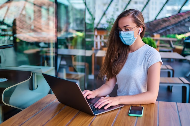 Casual working woman freelancer in protective mask, white wireless headphones and eyeglasses remotely works at the computer in cafe. Health protection in public places