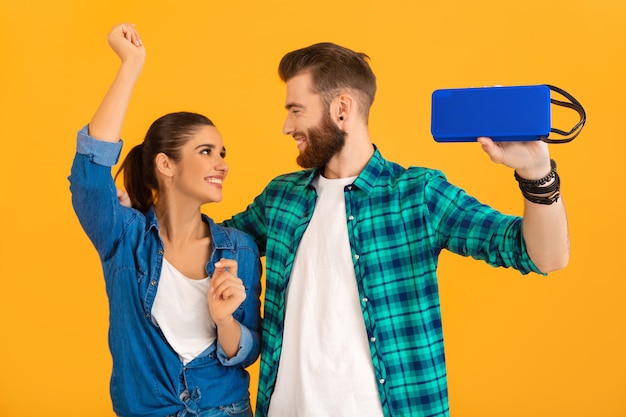 Casual young couple holding wireless speaker listening to music dancing isolated on yellow