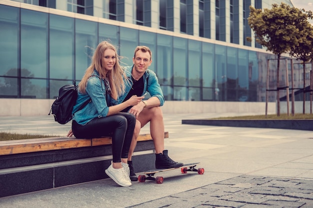 Casual young couple with smartphone and longboar over large modern glass building background.