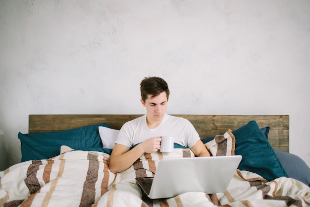 Casual young man using laptop in bed at home and drinking coffee