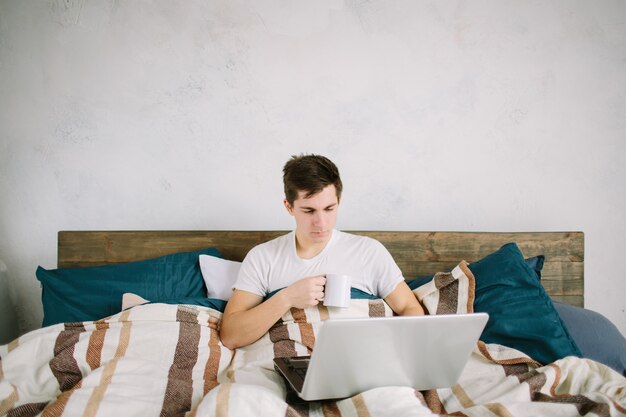 Casual young man using laptop in bed at home and drinking coffee