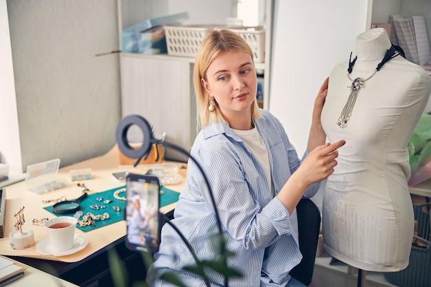 Casually dressed jeweler presenting trendy necklace on mannequin while recording product presentation with smartphone