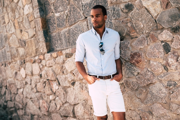 Casually handsome. Handsome young African man in smart casual clothes holding hands in pockets and looking away while standing against the stoned wall outdoors