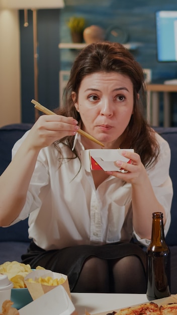 Caucasian adult looking at TV while eating chinese food and drinking beer from bottle sitting on living room sofa. Young woman enjoying asian meal and beverage at home after work