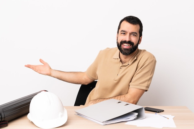 Caucasian architect man with beard in a table extending hands to the side for inviting to come.