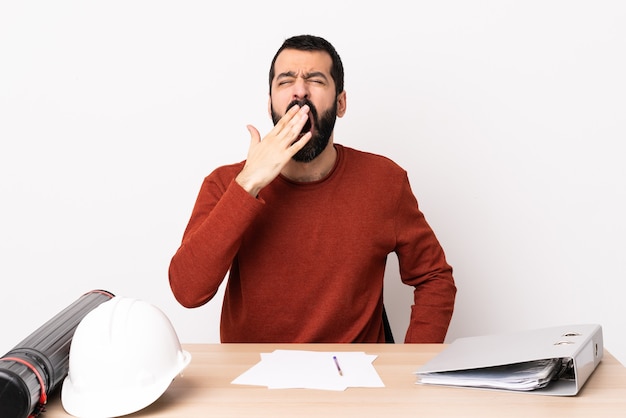 Caucasian architect man with beard in a table yawning and covering wide open mouth with hand.