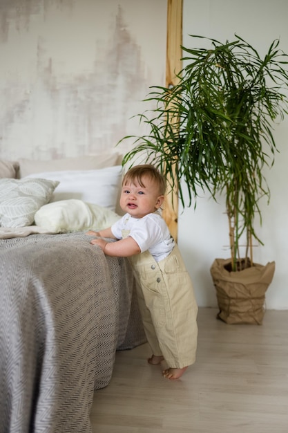 Caucasian baby boy in beige overalls is standing on the floor near the bed in the room The kid learns to walk around the bed