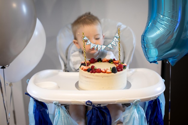 Caucasian baby boy celebrating first birthday at home Birthday berry cake Blue decoration balloon