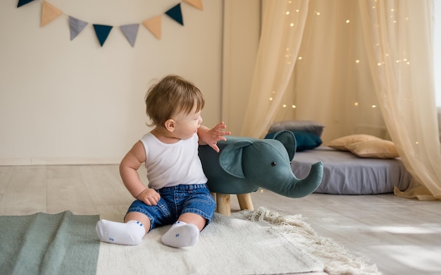 Caucasian baby boy is sitting on the floor in the nursery and playing with a soft elephant toy Playful baby boy in white bodysuit and jeans