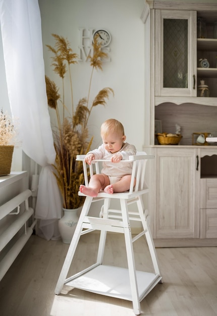 Caucasian baby girl in a white cotton bodysuit sitting in a wooden chair in the kitchen