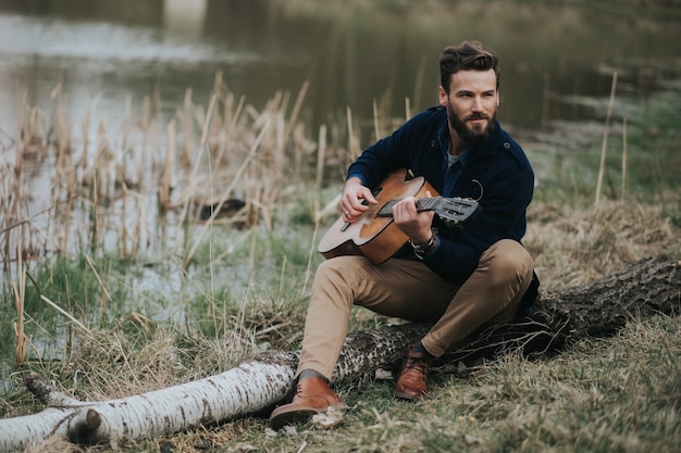 Caucasian bearded man is playing guitar near the lake. handsome stylish hipster.