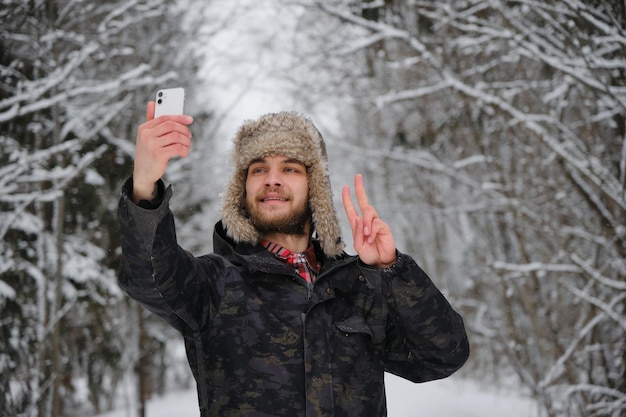 Caucasian bearded young handsome man wears fluffy hat with earflaps and takes selfie on phone