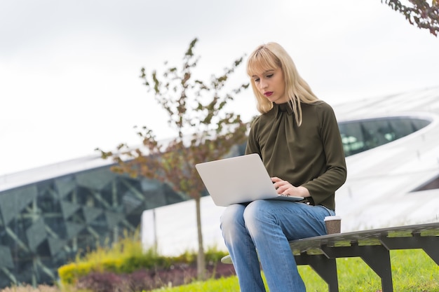 Caucasian blonde girl entrepreneur business park sending an email with laptop. Modern polygon among nature