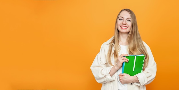 Caucasian blonde student girl holding a book notebook smiling looking at the camera isolated over orange background in studio Copy space