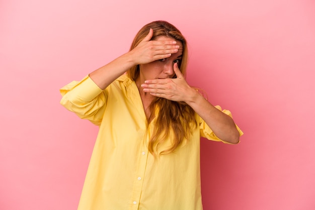 Caucasian blonde woman isolated on pink background blink at the camera through fingers, embarrassed covering face.