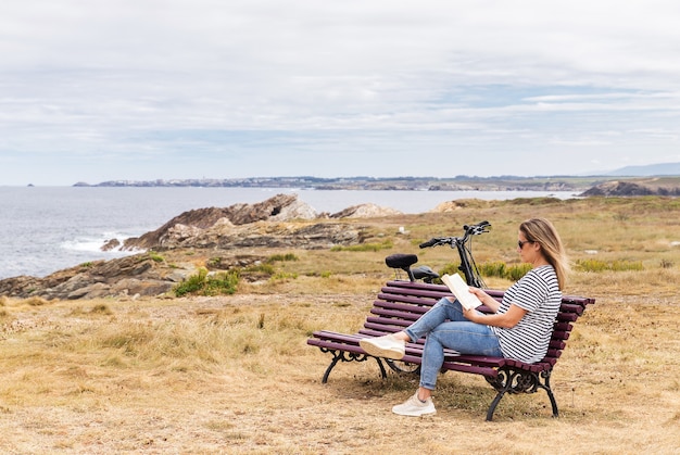 Caucasian blonde woman sitting on a bench in front of the sea reading a book