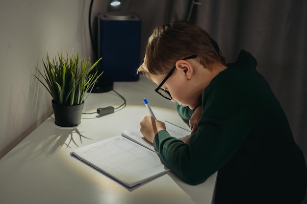 Caucasian boy doing homework at home in dark room by the light of lamp