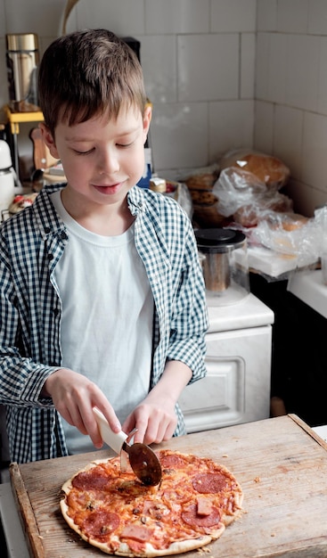 A caucasian boy in a shirt cuts pizza with salami with a round knife in the kitchen on the table round pizza with tomatoes and salami on a wooden backgroundItalian fast food