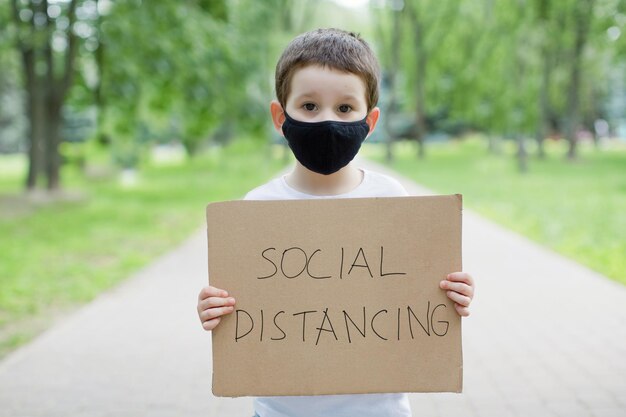 Caucasian boy with social distancing carboard sign Child in black facemask with cardboard sign outdoor