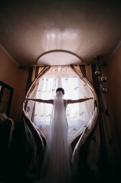 caucasian bride in a wedding dress opens the curtains on the window in the bedroom, wide angle photo