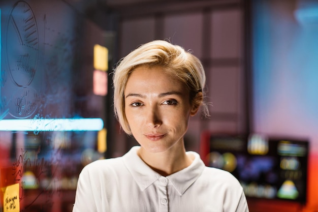 Caucasian business woman in white shirt posing on camera while standing in modern dark office