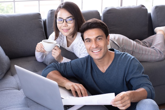 Caucasian businessman and Asian businesswoman wearing casual dress sitting together in living room and working on laptop notebook computer with relax and happiness. Idea for modern working at home