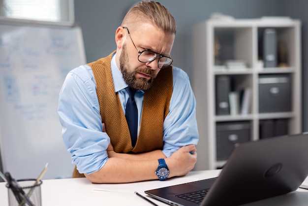 Caucasian businessman in eyeglasses sitting at desk with modern laptop and looking on screen with serious facial expression Concept of people occupation and technology