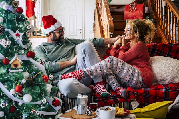 Caucasian couple having fun while sitting on sofa in living room during Christmas celebration at home. Couple in hat and socks with decorated Christmas tree.