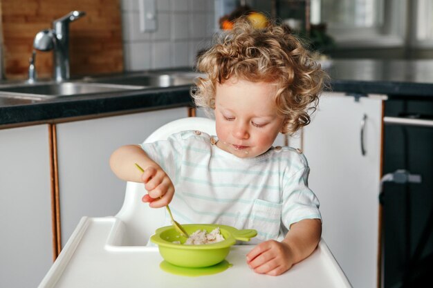 Caucasian curly kid boy sitting in high chair eating cereal puree with spoon healthy eating