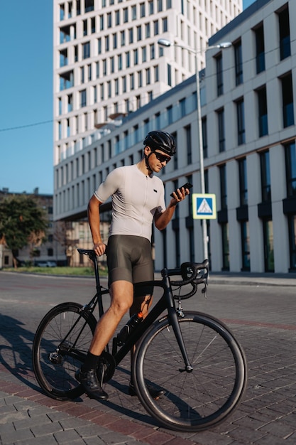 Caucasian cyclist in sportswear looking on mobile screen while standing on city street with black bike Man taking break during workout outdoors